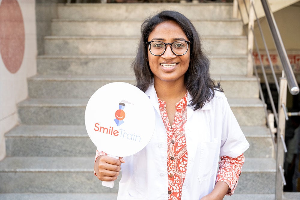La Dre Mayuri en sarrau de laboratoire tenant une affiche de Smile Train