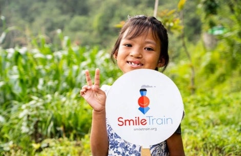 Smile Train patient smiling, flashing a peace sign, and holding a Smile Train sign after cleft surgery